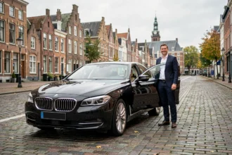 Chauffeur VTC professionnel en costume sombre debout près d'une berline noire sur un boulevard pavé de Lille avec architecture flamande en arrière-plan
